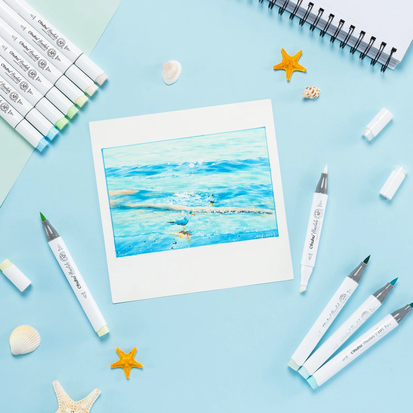 Artistic watercolor painting of a blue ocean scene on a white card, surrounded by art supplies on a light blue background.