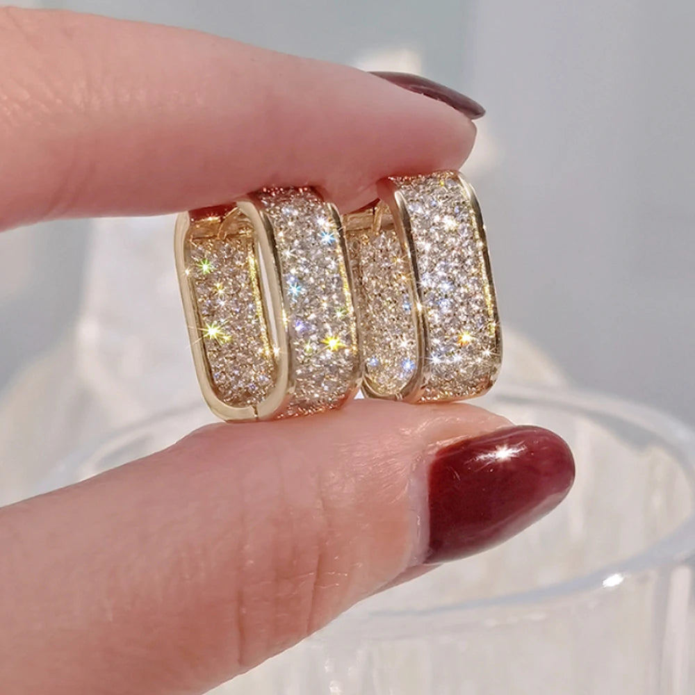 Gold hoop earrings with sparkling stones held between fingers against a neutral background