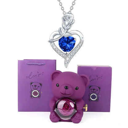 Purple teddy bear with rose, silver necklace with blue heart pendant, and purple packaging on white background
