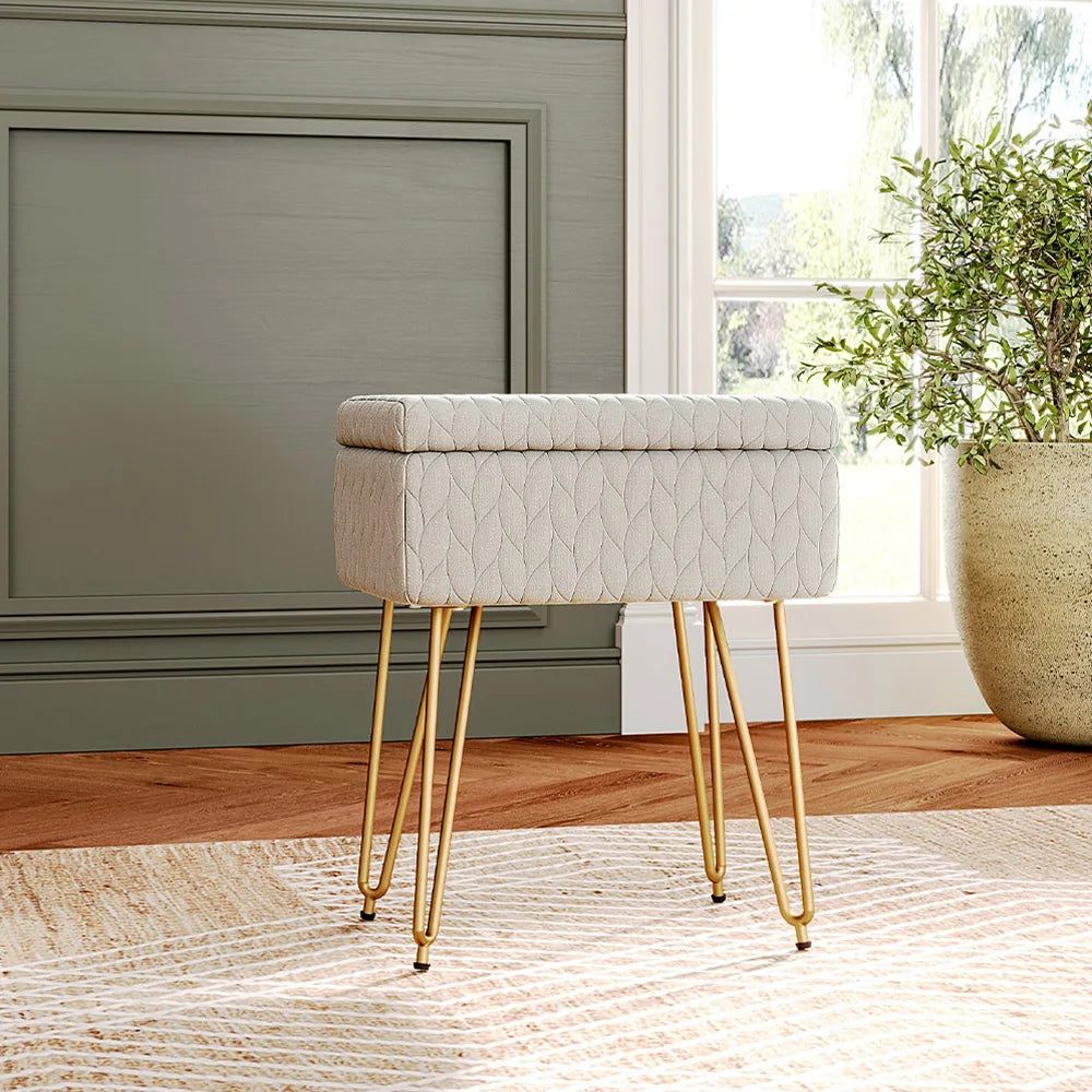 Decorative stool with gold legs and textured beige cushion in a room with a window and plant.