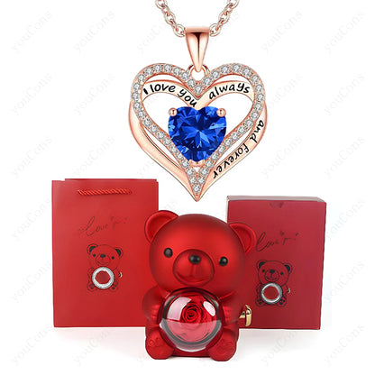 Heart-shaped necklace with blue gem and rose gold accents, red teddy bear with rose, and red packaging on a white background.