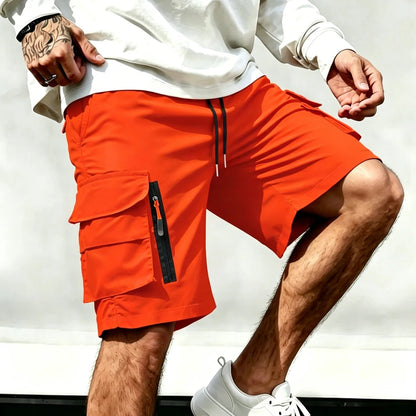 A man wearing bright orange cargo shorts with a black drawstring and a vertical zippered pocket on the left leg, styled with a white sweatshirt and white sneakers.