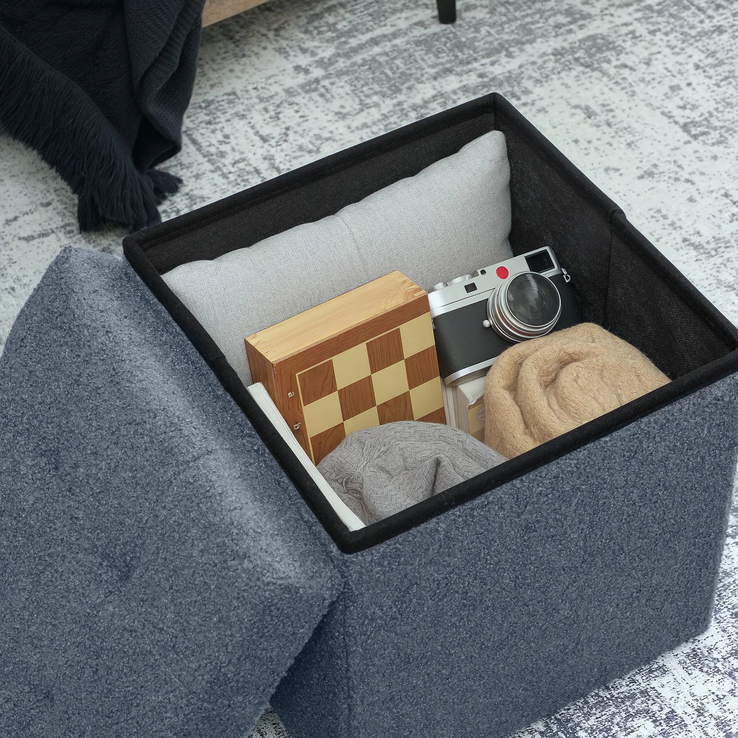 Gray storage ottoman with items like a camera and checkered box inside.
