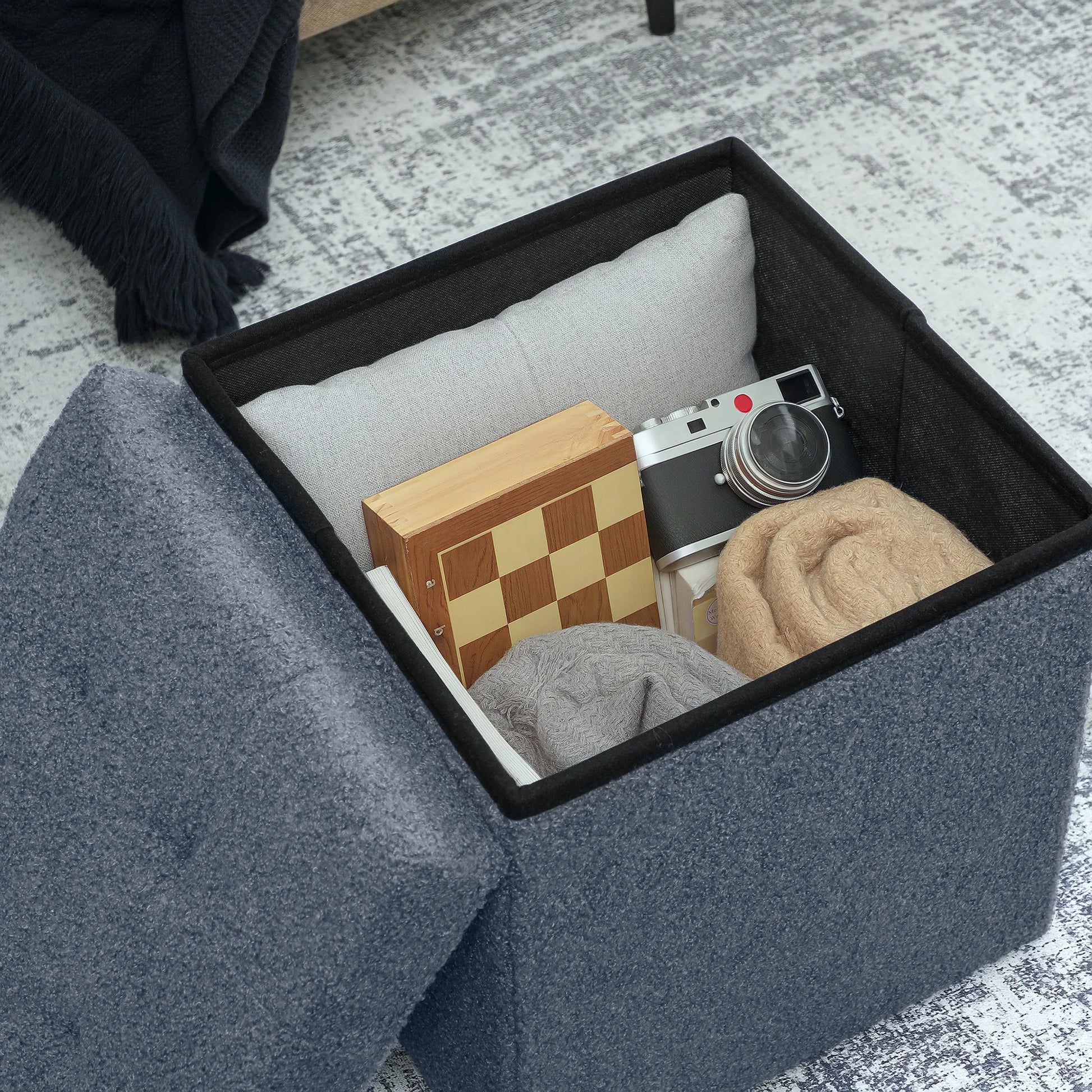 Gray storage ottoman with items like a camera and checkered box inside.