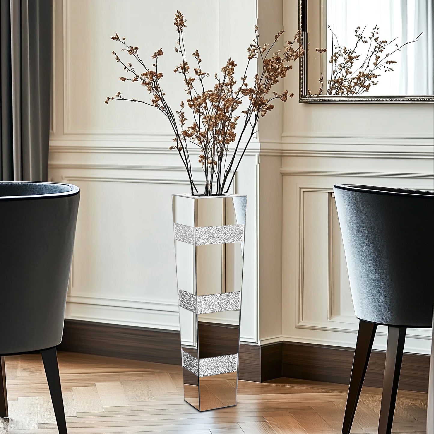 Tall silver vase with decorative branches in a room with two black chairs and a mirror.