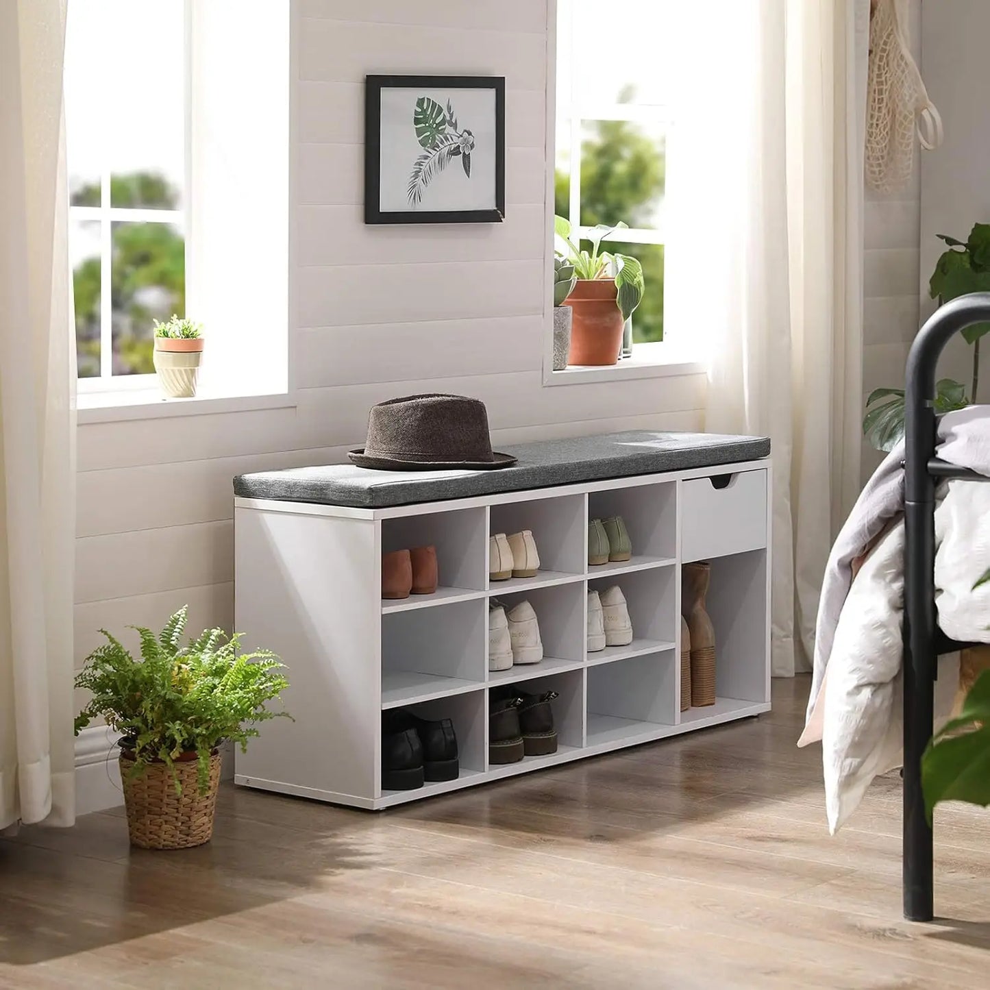 White shoe storage bench with black top in a room with plants and a bed.